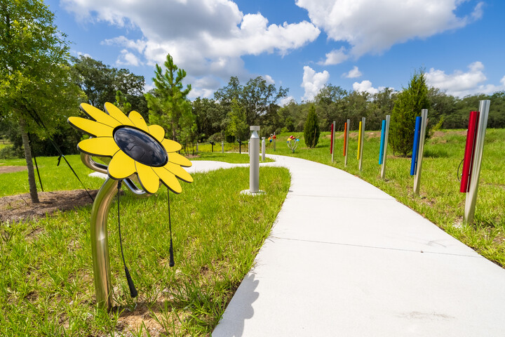 Sunflower-shaped sculpture and colorful vertical chimes along a winding pathway through a landscaped outdoor space.