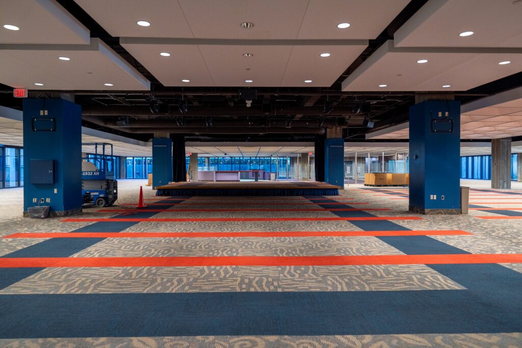 Large open interior with blue columns, striped carpet, and a temporary stage centered in the room during renovation or setup.