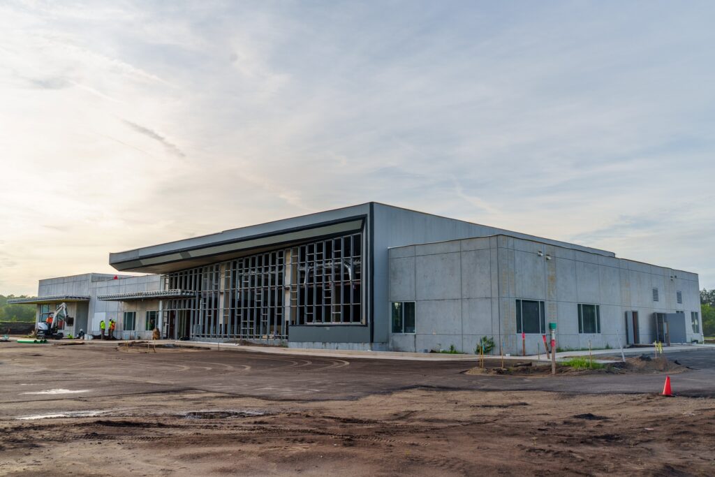 Large concrete building under construction beneath a cloudy sky, with workers and equipment near the unfinished entrance and surrounding grounds.