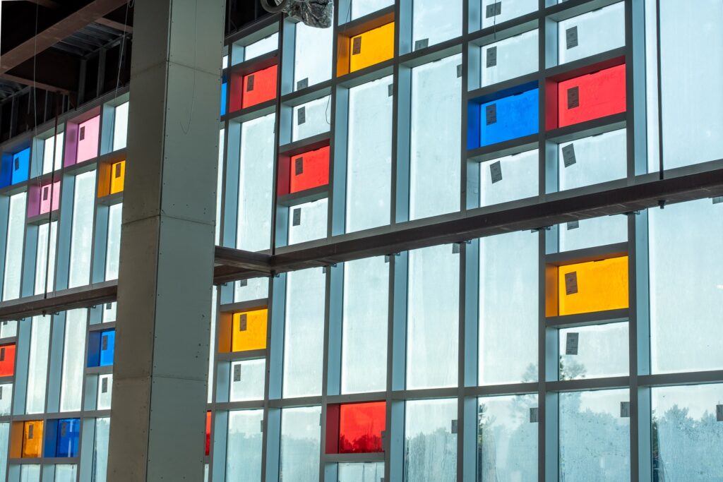 Floor-to-ceiling window wall inside a building under construction, featuring scattered red, blue, yellow, and pink glass panels among clear panes.
