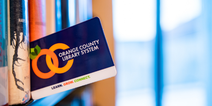 OCLS library card tucked between books on a shelf in front of a window