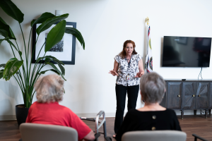 A presenter gestures while speaking to older adults seated in a bright community room.