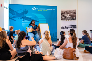 A storyteller holds up a colorful picture book for children and caregivers seated on the floor in front of an “Orlando Executive Airport” display and historic aviation photographs.