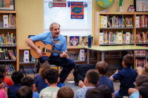 A library staff member plays an acoustic guitar for children seated on the floor in a brightly decorated library.