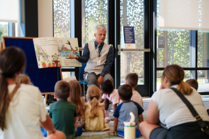 A seated library staff member displays a large illustrated book while presenting storytime to children and caregivers.