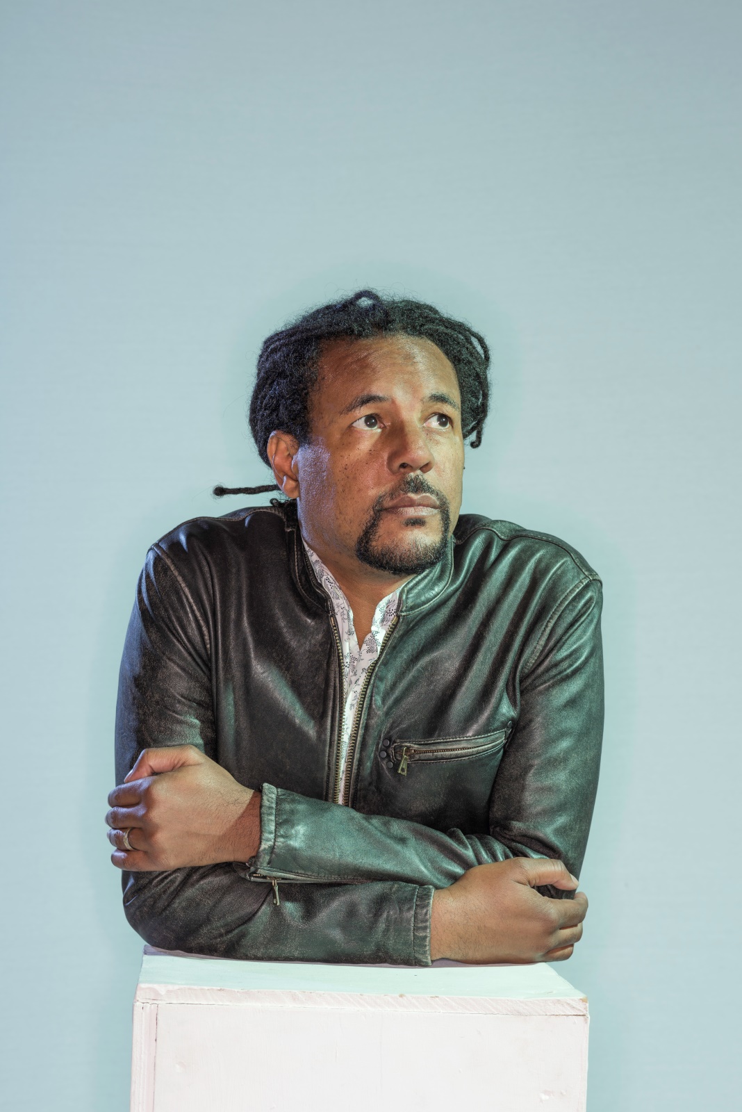 Author Colson Whitehead in a dark leather jacket, leaning forward with arms crossed against a light blue background