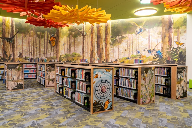 Children’s book area with woodland-themed shelves, a wraparound forest mural, and orange and yellow ceiling decorations.