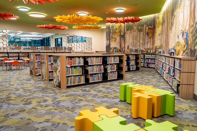 Children’s library with low wooden bookshelves, colorful puzzle-piece seating, honeycomb dividers, and a woodland mural.