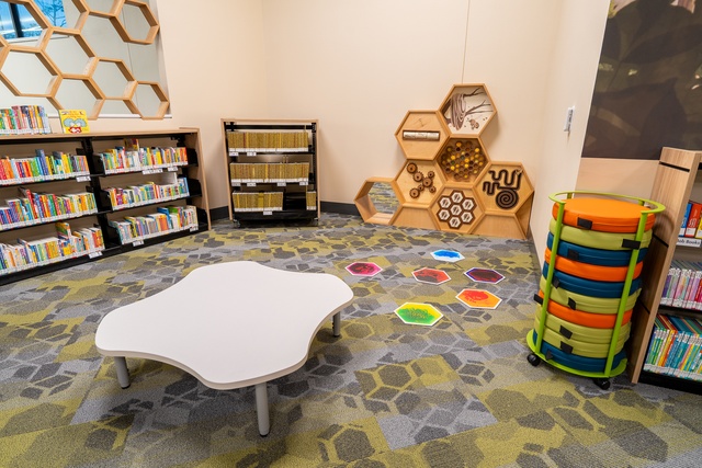 Children’s library activity area with a low white table, bookshelves, colorful floor markers, stacked stools, and honeycomb-shaped wall displays.